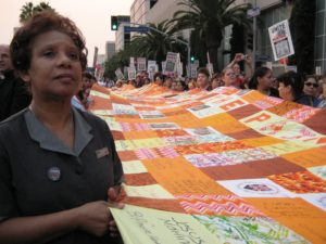 Workers stand closely together holding up an orange and yellow quilt. A brown-skinned woman in the foreground looks off into the distance. She is wearing a maid's uniform with a name tag and a UNITE-HERE button.