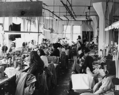 Black and white image of the interior of a garment shop in downtown LA. The workers, all women, sit at sewing machines, many of which have power cords extending above them to the ceiling. There is lots of surplus fabric around and florescent lights hanging with the cords from the machines. the work stations are numbered. One woman looks over her shoulder and catches the camera's eye.