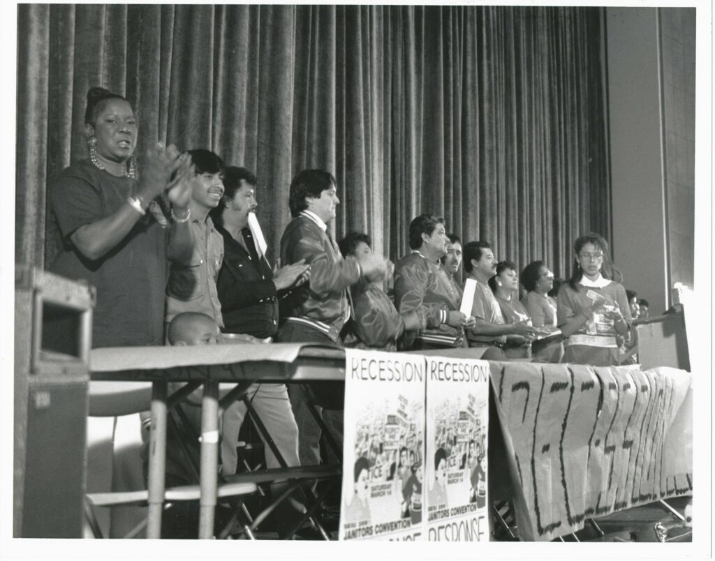 Black and White image of a stage with a dozen people standing at tables with banners on the front of them