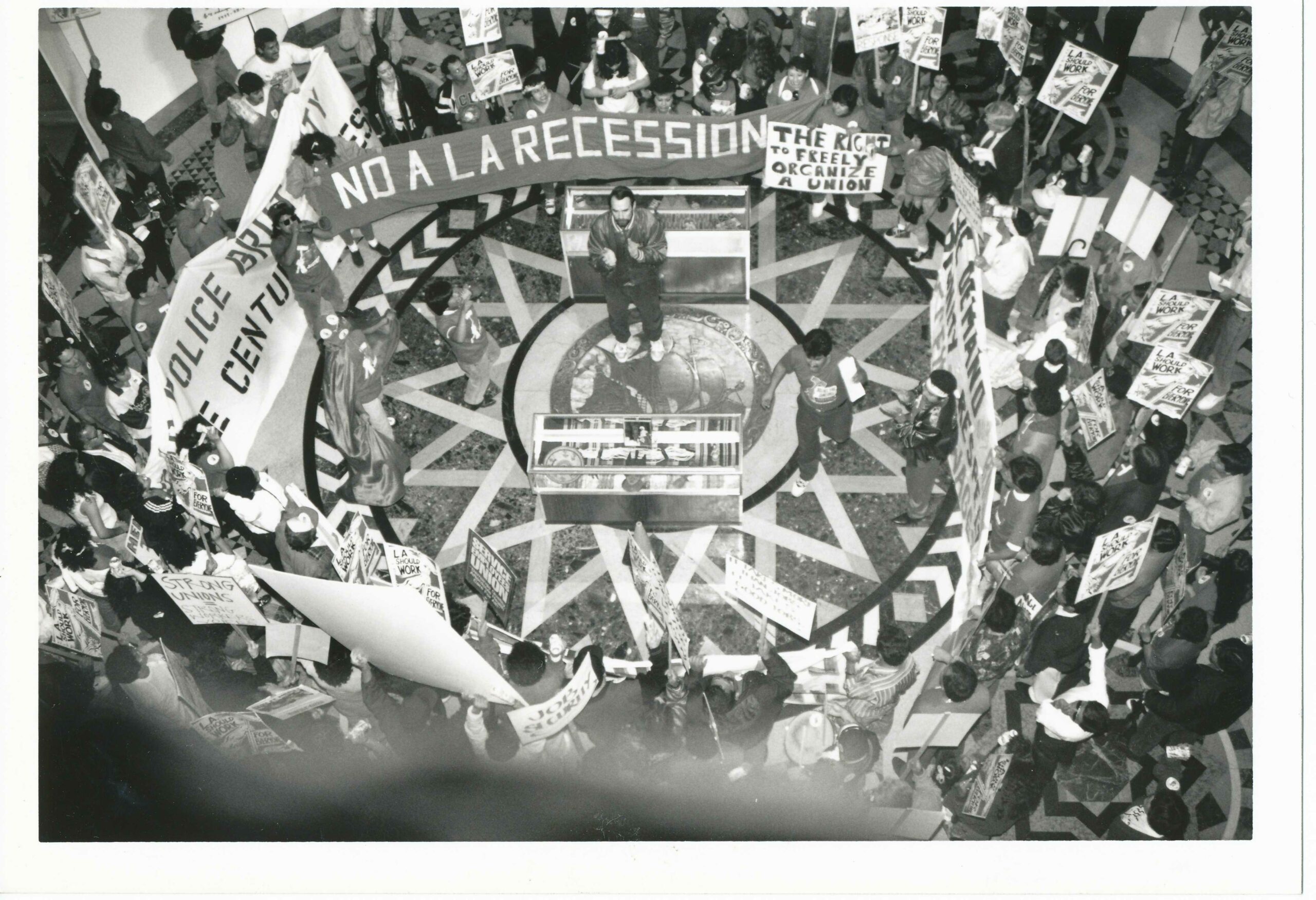 black and white image taken from above of a group of protestors carrying banners ("No a la Recession") and Picket Signs ("L.A. Should Work for Everyone") gathered in a circle in the rotunda of City Hall. In the center, a man wearing a silk jacket claps his hands.