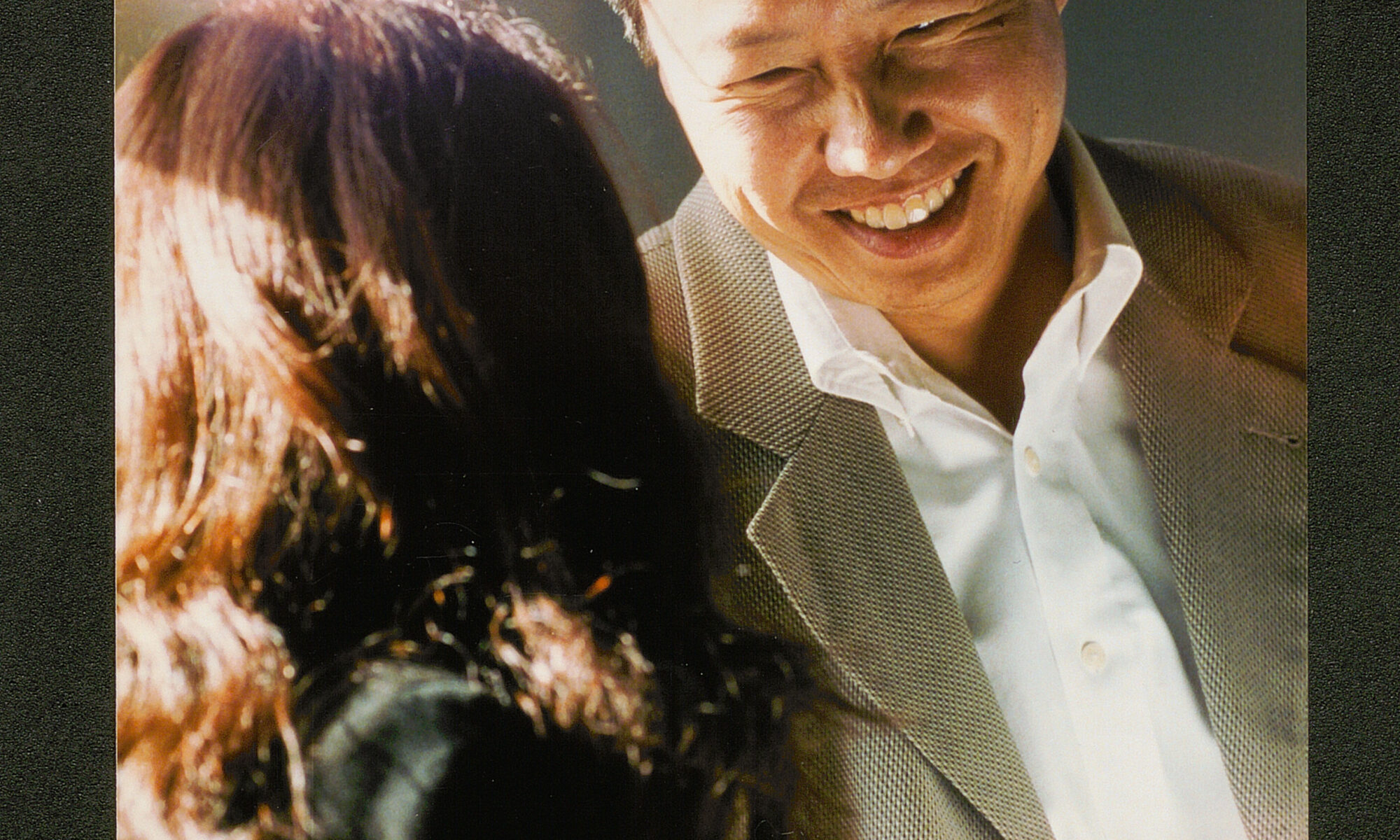 A tall Asian man in a tan suit with no tie smiles as he speaks with a shorter woman in a black blazer, her back faced to the camera. They are standing next to a window with the sunlight casting on their faces.