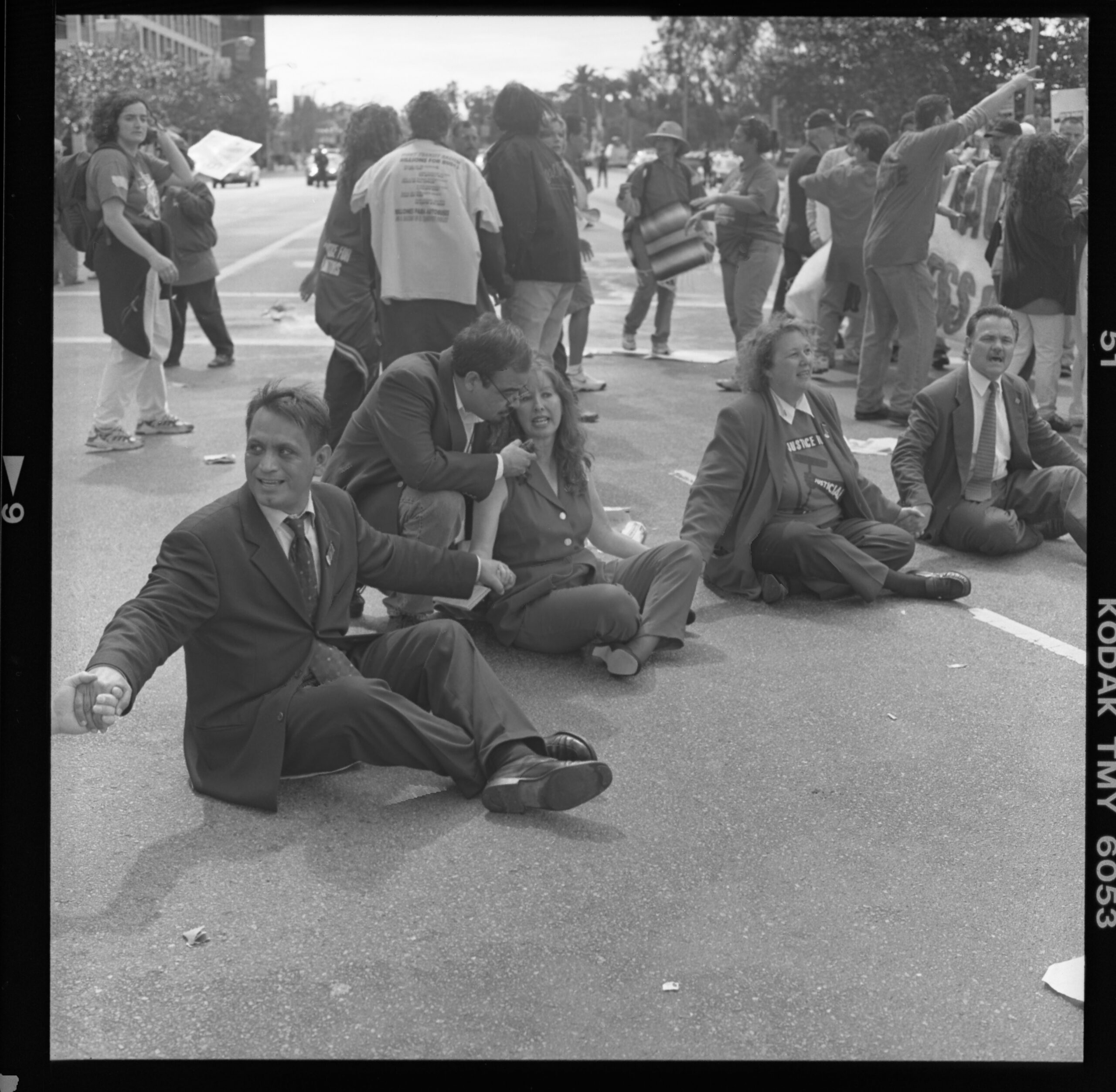 Black and white photograph of four people sitting in the street holding hands in a circle. The man on the left is in a suit and looks to his right. The woman next to him speaks with a reporter crouched to the ground. They are surrounded by protestors shouting and moving in various directions.