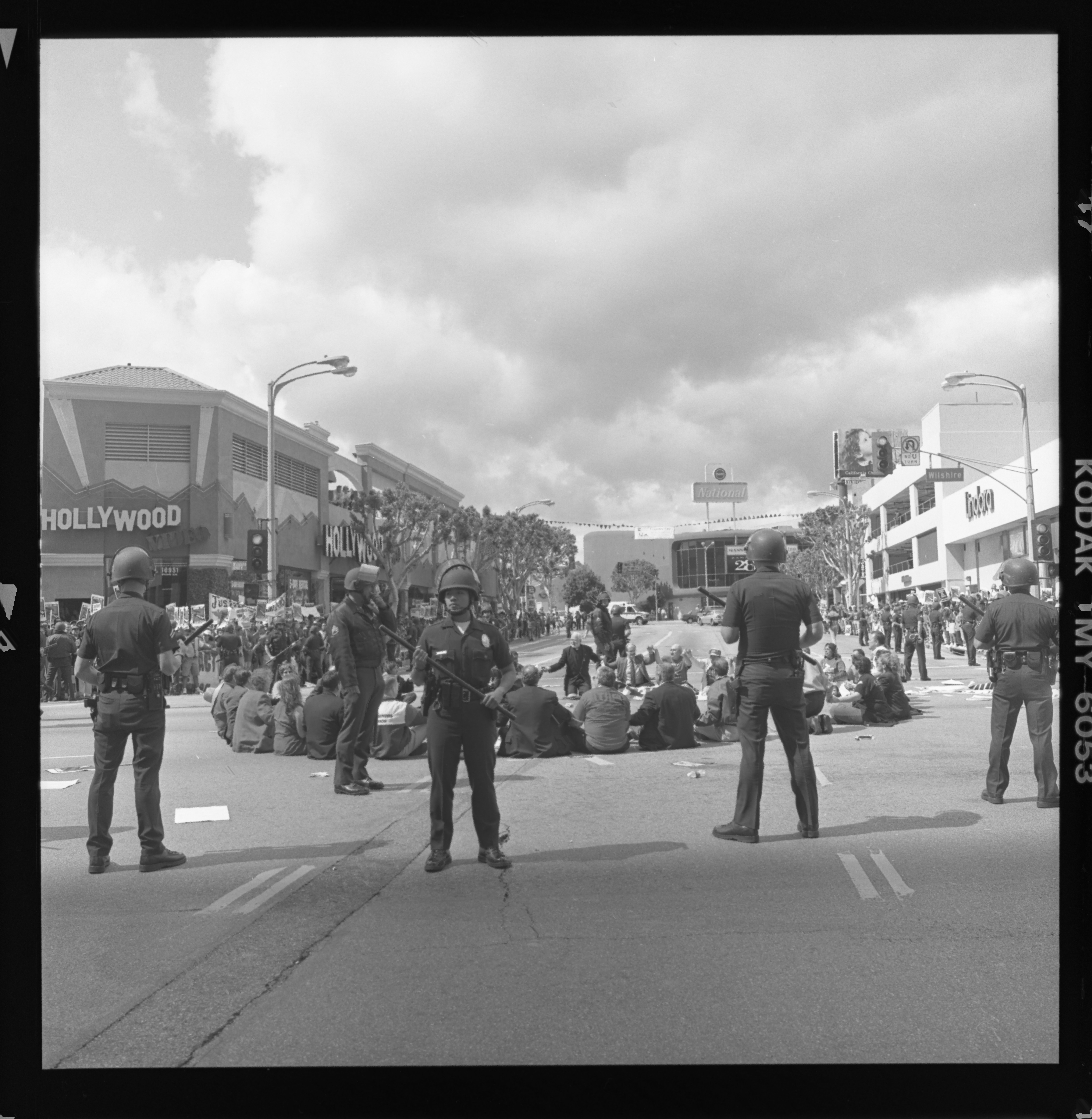 Striking street-level view of a group of people sitting in the middle of an intersection in a circle holding hands, most with their backs to the camera. In the foreground of the image are five police officers standing over them holding battons. Surrounding them on the sidewalks are protestors carrying banners and picket signs. There is a street sign visible that reads "Wilshire"