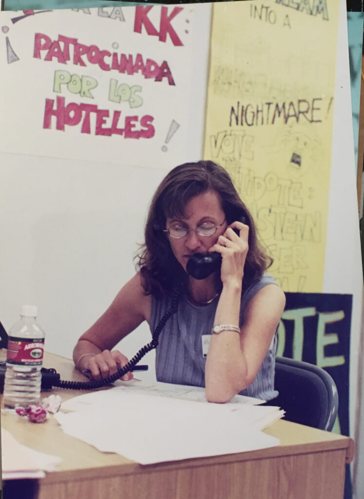 A brown-haired woman (Madeline Janis) sits at a desk making a phone call. Behind her are signs reading "KK: Patrocinada for los hoteles!" and "Nightmare!"