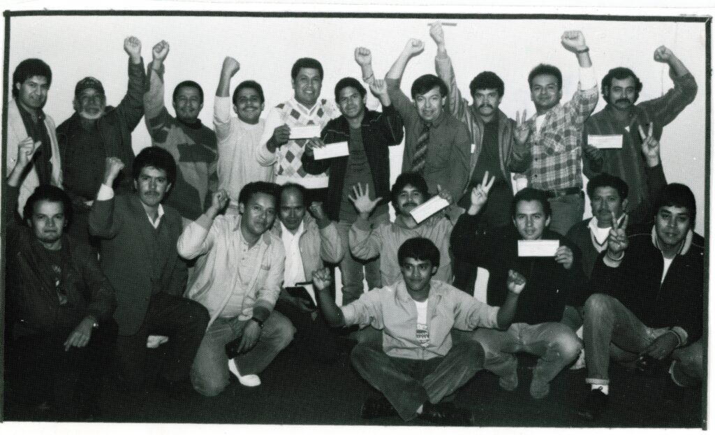 A group of men raise their fists and make victory signs. Some hold paper checks.