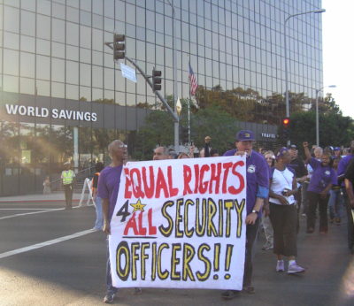 Workers in purple t-shirts carry a banner reading "Equal rights 4 All Security Officers!!" lead a crowd of demonstrators marching in the street. Behind them is a glass office tour with a "World Savings" logo. A police officer directs traffic. Sunbeams break around the edge of the left hand side of the frame.