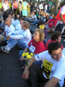 A woman in a red shirt sits crosslegged into in street surrounded by others doing the same. Her expression is calm but determined.