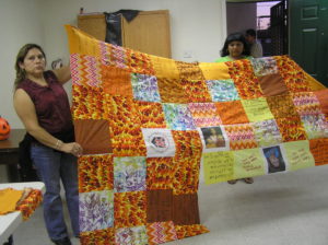Two women hold up a quilt in production made of mostly orange and yellow patches. Some of the patches have hand written messages on them.