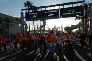 a group of marchers, lead by people wearing bright orange safety vests, pass under large street signs reading "Arriving Flights," "Departing Flights," and "To Highway 1, Sepulveda Boulevard." behind the march (the end of which is not visible) looms the decorative statues at the gateway to LAX.