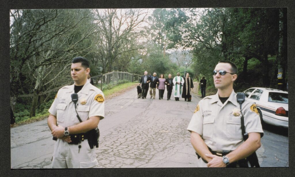 scene of a quite street lined by oak trees. in the foreground, two police officers stand facing the camera. In the background, 6 clergy members, most wearing robes and/or collars, walk down the street towards the cops with their arms locked. They can be seen smiling. One of them walks a service dog.