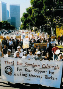 A grainy color photograph of a large demonstration with the downtown L.A. skyline in the background. At the front of the march, several people carry a large banner reading "Thank you Southern California For Your Support for Striking Grocery Workers."