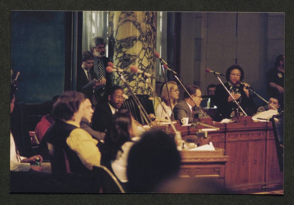 A photograph inside the chambers of the Los Angeles City Council, taken from the angle of the public seating. One councilwoman stands to address the chamber, while others sit at their desks.