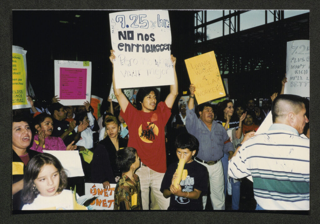 A crowd of people carrying homemade signs reading "Living Wage 4 LAX Workers" and "$7.25x hr no nos enrrianecero per vivid major." The man in the middle of the crowd wears a red union t-shirt and lifts his sign over his head.