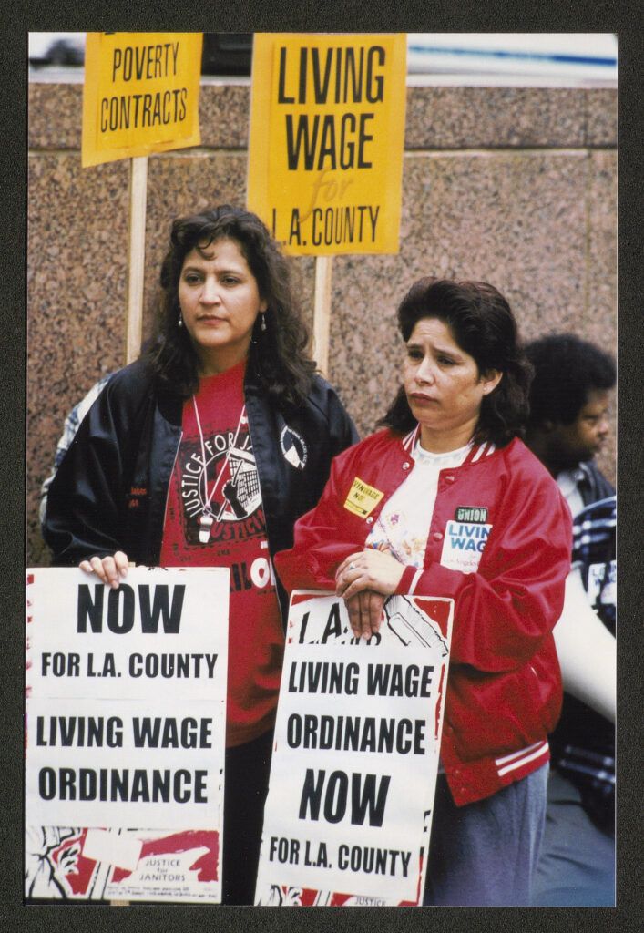 Two women holding picket signs wearing silk jackets and union t-shirts. Their signs read "Now for LA County - Living Wage Ordinance" and "Living Wage Ordinance Now for LA County"