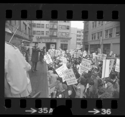 Black and white image appears as a negative, with perforated strips on top and bottom. It depicts a group of workers in an alley outside a garment factory with a visible marquee reading "Dresses." The workers carry signs reading "IBEW joins ILGWU to Keep Americans Working" and "Stop Exporting Jobs". Mariachi musicians play trumpets and violins for the crowd from a stage.