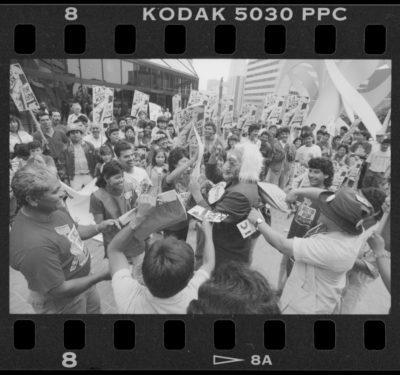 Black and white image of photo negative shows a crowd of workers, some wearing union t-shirts, gathered around a costumed figure in suit. They are wearing a full face mask of an old man with exaggerated features. The janitors laugh as they perform a street speaking skit.