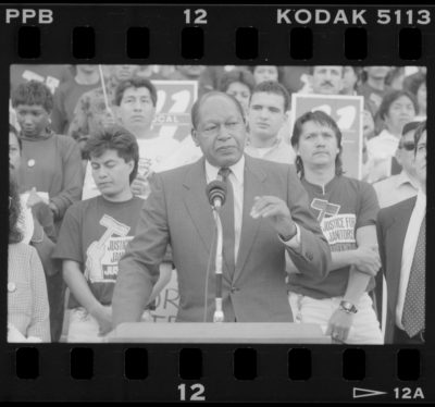 Black and white film negative of photograph of Mayor Tom Bradley speaking with members of the Justice for Janitors standing behind him. The two Latinx men standing directly behind Mayor Bradley are wearing union T-shirts with the Justice for Janitor's logo of a raised fist holding a mop.