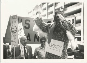 Black and white image of a person wearing a turkey suit (sign hung around their neck reading "turkey") at a protest. Protestors carry picket signs reading "Turkey of the Year - Jonel C. Hill." One older white man wears a suit and tie with a Justice for Janitors pin.
