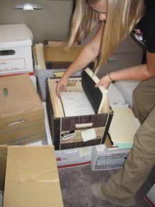 a blonde woman (face out of frame) shows the contents of a banker's box in a dusty room.