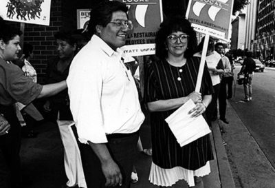 Low-resolution black and white image of two smiling people, Miguel Contreras and Maria Elena Durazo, near a Local 11 picket line. Durazo is holding an HERE Local 11 picket sign.
