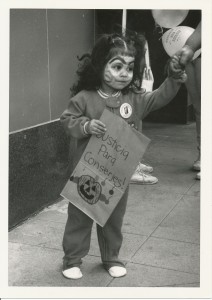 black and white image of a very cute little girl in a pajama romper with footies. Her face is painted for halloween and she carries a brown bag with a jack-o-lattern drawn on it that says "Justice para Conserjers!" An adult holds her hand and balloons can be seen behind her.
