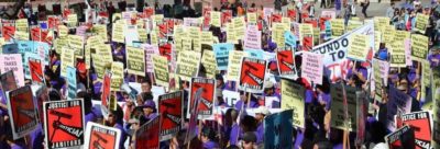 a sea of picket signs being held up by workers wearing purple t-shirts. The signs read "The 1% is holding us ALL BACK," "We are all the 99%," "The 1% Takes $9,200 from your Middle Class Family," and Justice for Janitors."