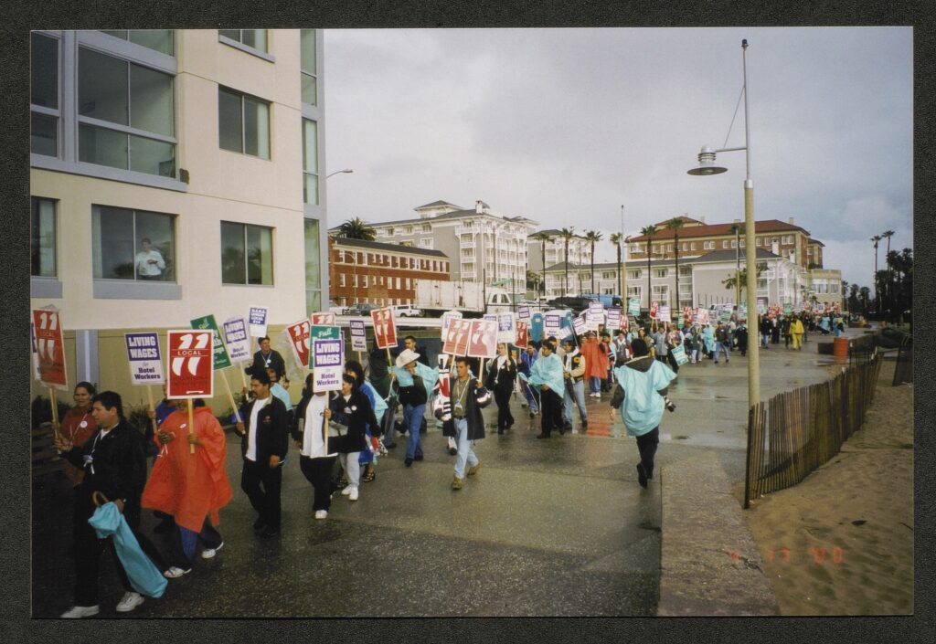 A procession of workers wearing raincoats carrying pickets marching down the Santa Monica boardwalk. Some carry red signs reading "Local 11," Purple and white signs reading "Living Wages for Hotel Workers" and green signs reading "Full Family Health Coverage." A man can be seen working through the window of one of the hotels in the foreground. The pavement appears wet and the sky looks as if it has just rained.