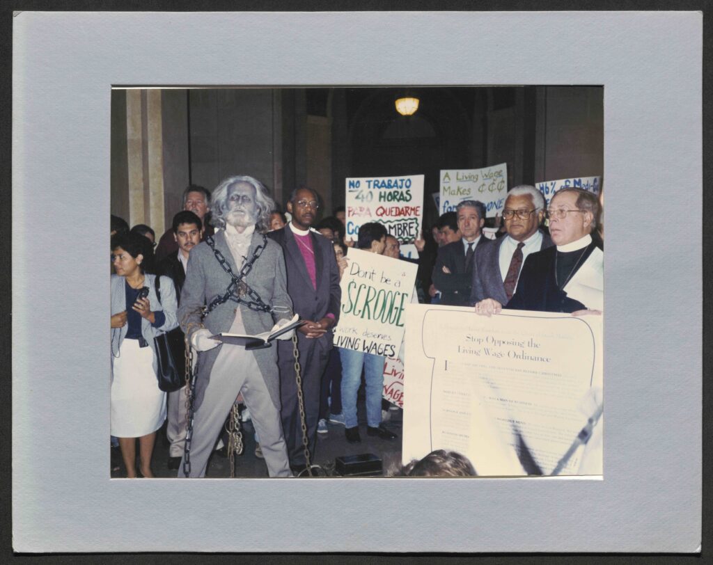 a crowd of religious leaders and workers gathered in Los Angeles City hall. On the right, a priest in his white collar and black suit holds a large sign reading "Stop Opposing the Living Wage Ordinance." On the left, a man stands wearing a costume, make up and a wig, posing while reading from a large book in his hands. He is dressed as the ghost of Jacob marley, wrapped in chains. In the background of the image, people hold signs reading "No Trabajo 40 Horas Para Quedarme...[illegible]" "Don't be a Scrooge, work deserves living wages," and other messages.