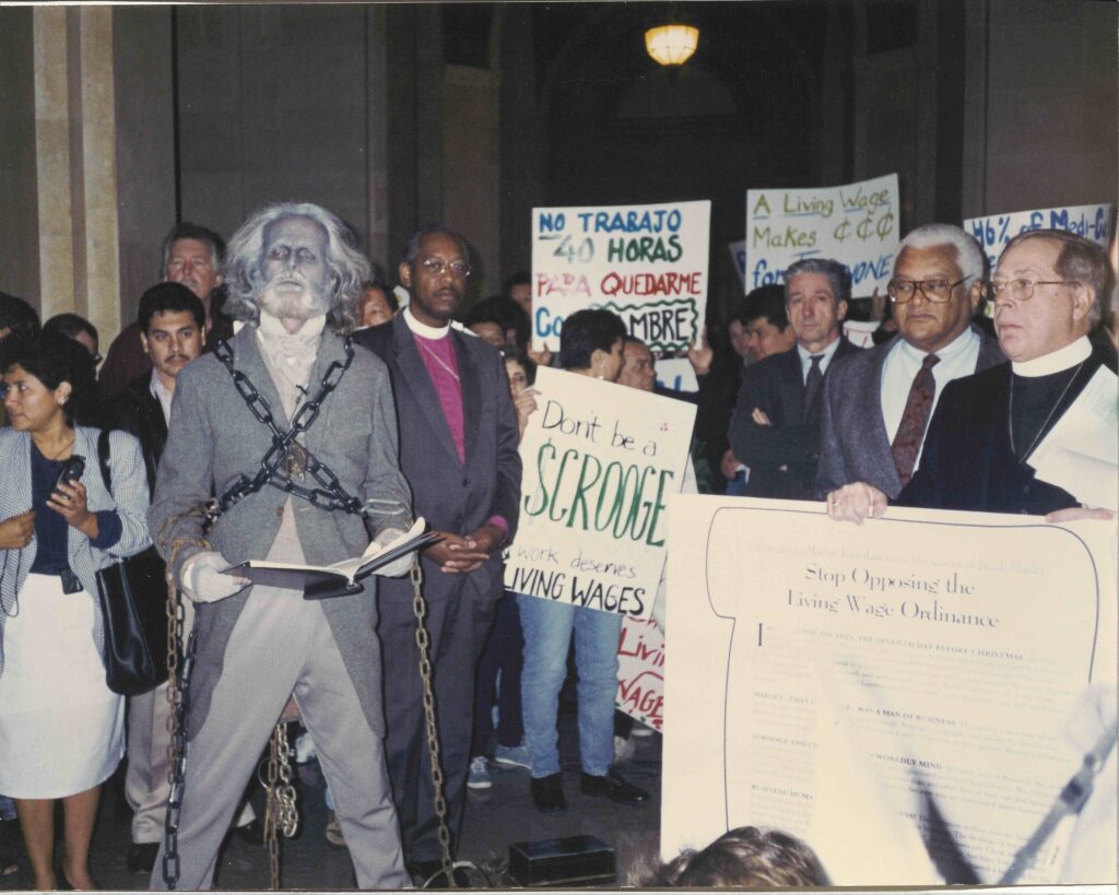 A frame color photograph of a man dressed as Jacob Marley surrounded by workers and religious leaders (in collars).