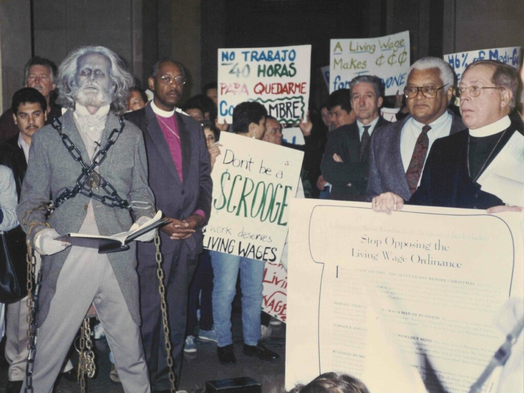 a crowd of religious leaders and workers gathered in Los Angeles City hall. On the right, a priest in his white collar and black suit holds a large sign reading "Stop Opposing the Living Wage Ordinance." On the left, a man stands wearing a costume, make up and a wig, posing while reading from a large book in his hands. He is dressed as the ghost of Jacob marley, wrapped in chains. In the background of the image, people hold signs reading "No Trabajo 40 Horas Para Quedarme...[illegible]" "Don't be a Scrooge, work deserves living wages," and other messages.