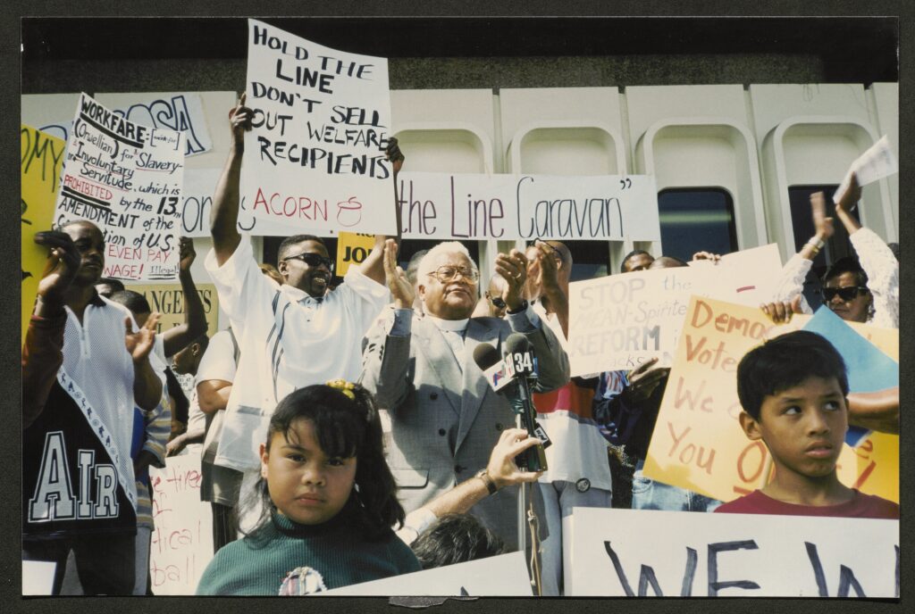 A crowd carrying home made signs reading "Hold the Line - Don't Sell out Welfare Recipients - ACORN" and other messages. In the center of the crowd, an older priest wearing a white collar claps his hands while standing at a microphone. In the foreground are two children, one looking directly into the camera lens.