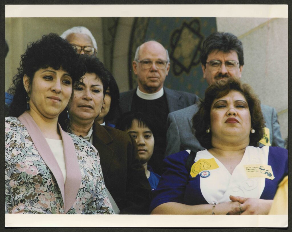 Two rows of people stand facing the camera. In the front row are three women dressed in business attire, one of whom has stickers fixed to her blue and white blouse reading "Local 11" and "Do the Right Thing!" as well as a blue, red, and white Local 11 pin. Behind them stand three men, all with glasses. the man in the center, framed by two of the women in front of him, wears a priest's white collar and black sweater and a blazer.