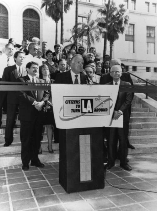 Black and white image of Mayor Richard Riordan standing at a podium and the bottom of the steps to LA City Hall. The podium bears a sign that reads "Citizens to Turn LA Around." He is flanked by other city council members and supporters, most of them men. 