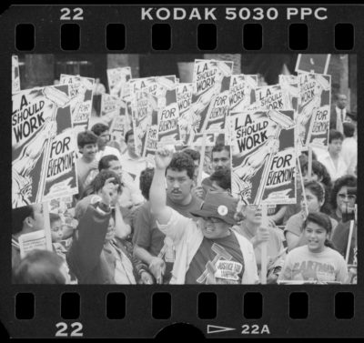 Black and white photo negative of a crowd of workers holding picket signs, which reads "L.A. Shoulw Work for Everyone" and features a cartoon strong arm raising a mop into the air.