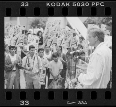 In the foreground, Jim Zellers speaks into a microphone while holding a picket sign. In the background, workers stand holding the same picket sign, which reads "L.A. Shoulw Work for Everyone" and features a cartoon strong arm raising a mop into the air.